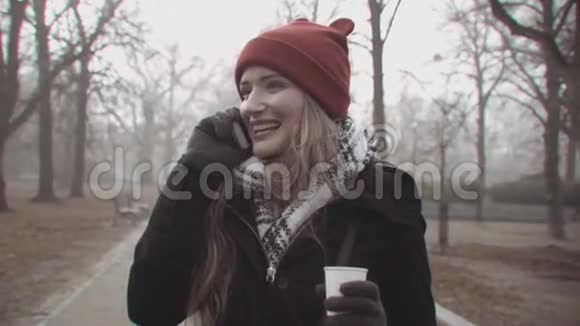 戴着红帽子的开朗女人拿着咖啡在户外打电话视频的预览图