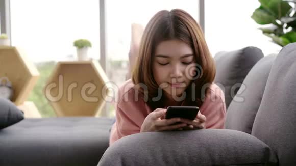 亚洲女人在客厅里躺在家里的沙发上用智能手机视频的预览图