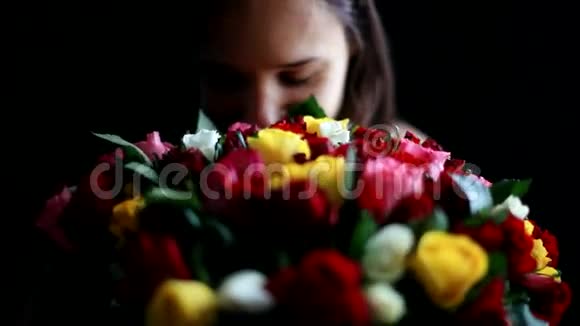 美丽的年轻女子怀里抱着一大束花闻着一朵芬芳的五颜六色的玫瑰背景是黑色的视频的预览图