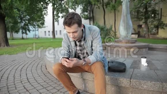 穿夹克衫的年轻人手里拿着电话坐在花岗岩圆面上夏季视频的预览图