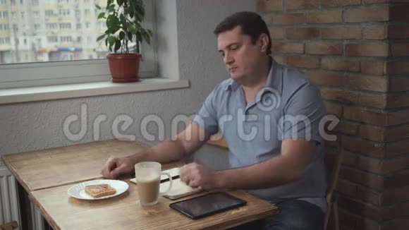 早上穿着蓝色衬衫的成年男性坐在咖啡馆里喝咖啡吃烤面包在桌子上和平板上视频的预览图