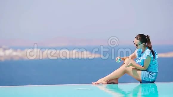 苗条的女人在她的腿上涂防晒霜坐在游泳池背景的边缘欧洲的老城Mykonos视频的预览图