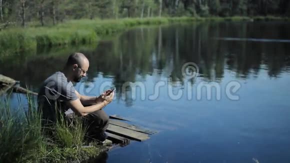 一个男人坐在湖边的码头上在电话里拍照背景上美丽的蓝色湖泊和森林视频的预览图