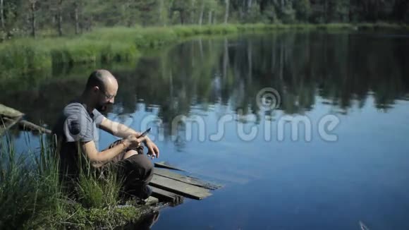 一个男人坐在湖边的码头上正在用视频打电话背景上美丽的蓝色湖泊和森林视频的预览图