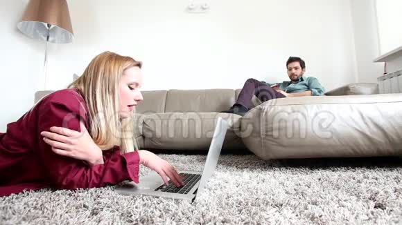 女人躺在地毯上看着笔记本电脑男人坐在背景沙发上视频的预览图