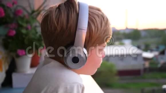 一个年轻人站在窗户附近戴上耳机听音乐背景模糊日落少年视频的预览图