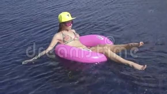 漂亮的年轻女人穿着比基尼黄色的帽子和紫色的太阳镜坐在游泳池里的粉红色充气戒指上视频的预览图