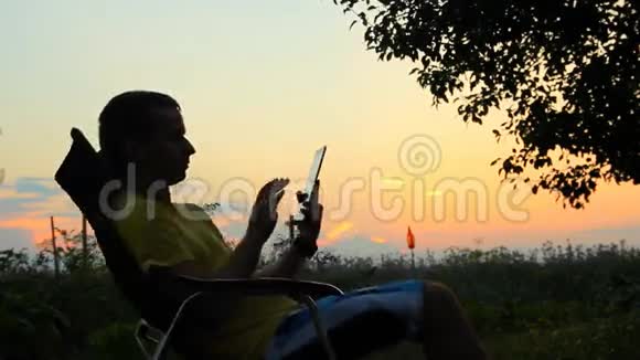 男性在椅子上玩平板电脑的剪影在日落后橙色天空的背景下视频的预览图