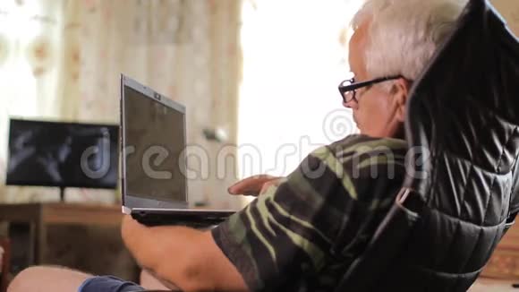 老人坐在靠近家庭电视的皮椅上手提电脑带着电脑放松的男人视频的预览图