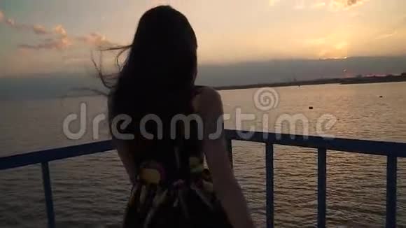 日落时穿着一件浅色衣服的漂亮女孩的剪影一个女孩正在海上走码头视频的预览图
