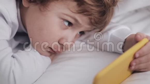 小男孩躺在床上饶有兴趣地看着现代黄色智能手机上的卡通视频的预览图
