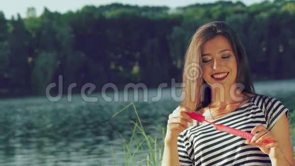 快乐的黑发在池塘附近的夏季公园里吹着五颜六色的肥皂泡慢慢地视频的预览图