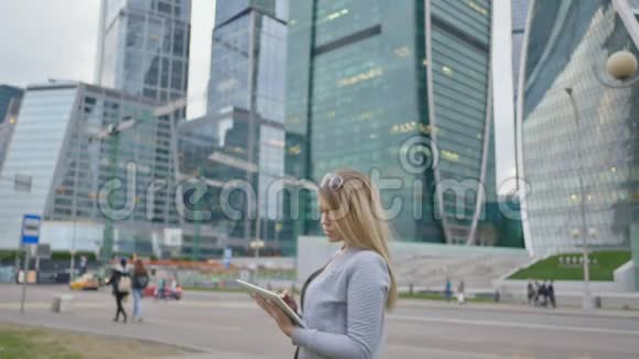 一个年轻的金发女孩在市中心摩天大楼的背景上使用平板电脑视频的预览图