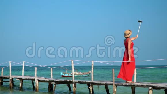 穿红色连衣裙和帽子的女人在Selfie棍子上拍照那个女人拿着Selfie棍子走在码头上慢慢视频的预览图