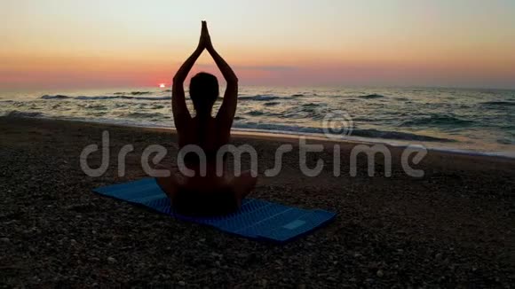 美丽的年轻女子坐在海滩上做瑜伽一边看日落视频的预览图