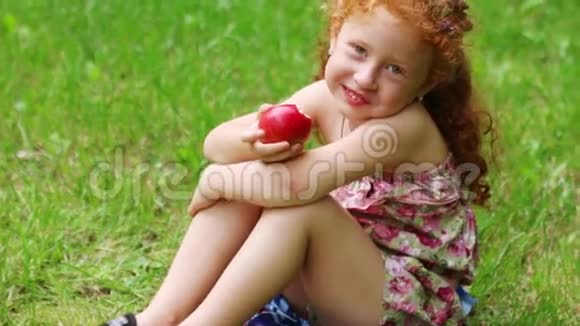 快乐的小女孩在绿色夏日公园的草坪上吃苹果视频的预览图