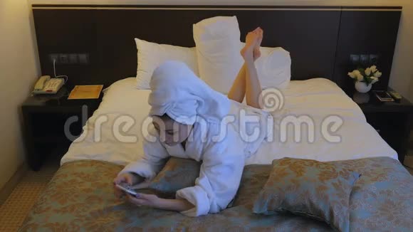 一个穿着白色外套的年轻漂亮的女人坐在酒店卧室的床上在电话里写短信视频的预览图