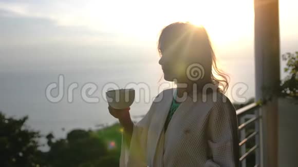 年轻漂亮的女人穿着浴袍在早晨喝着咖啡在美丽的日出站在她的露台上视频的预览图