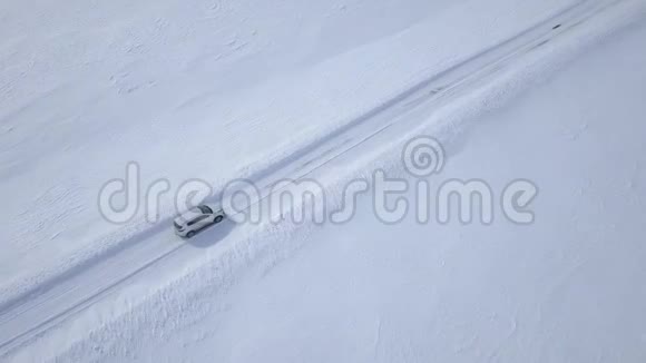 在白雪覆盖的田野之间的冬季森林道路上行驶的汽车的鸟瞰图视频的预览图