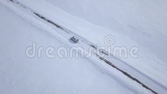 在白雪覆盖的田野之间的冬季森林道路上行驶的汽车的鸟瞰图视频的预览图