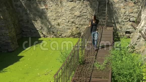 年轻漂亮的女孩走在被毁坏的生锈的金属桥上在绿水上视频的预览图