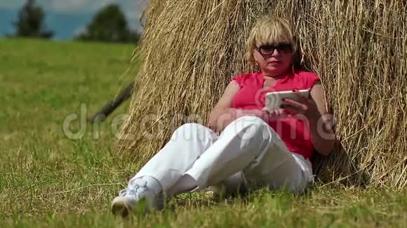 金发女人坐在干草堆附近的绿草上在他的智能手机上拍照视频的预览图