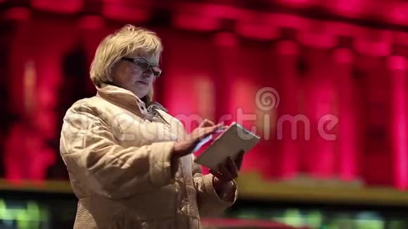有平板电脑的老妇人在夜间站在道路和红色建筑附近视频的预览图