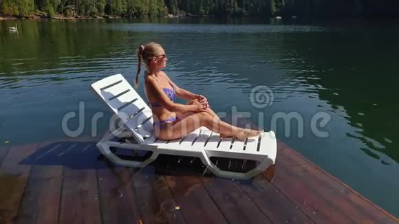 女人戴着太阳镜和泳衣坐在日光浴床上女孩在洪水木水下码头休息路面是视频的预览图