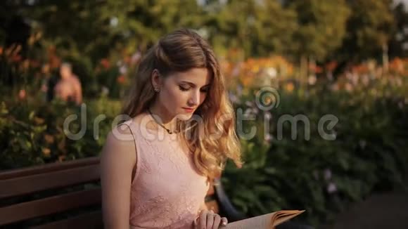 美丽的金发女郎坐在夏日公园的长凳上看书视频的预览图