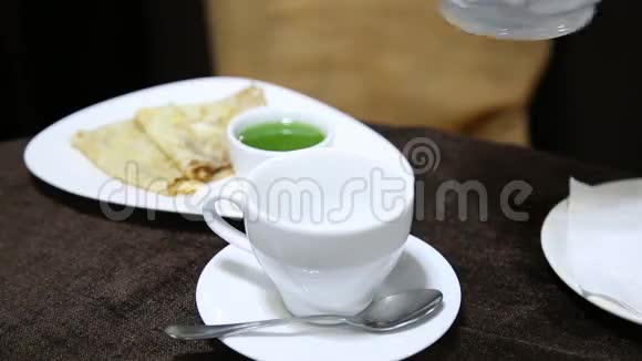 用瓷壶把茶倒入桌上的白杯中背景上的果冻和煎饼视频的预览图