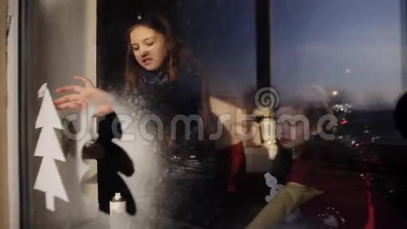 两个小女孩借助人造雪装饰一扇窗户为新年和圣诞节做准备视频的预览图