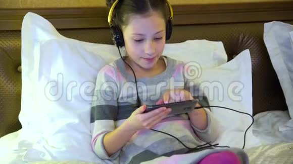 漂亮的小女孩坐在酒店的床上戴着耳机在智能手机上玩游戏视频的预览图