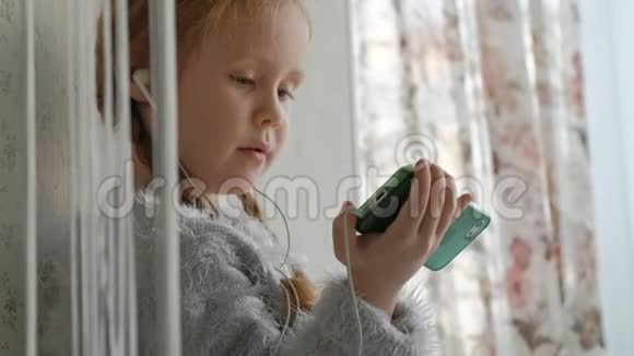戴着耳机梳着辫子的快乐小女孩看着手机视频微笑着坐在窗台上的房间里视频的预览图