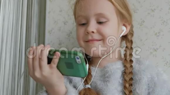 戴着耳机梳着辫子的快乐小女孩看着手机视频微笑着坐在窗台上的房间里视频的预览图
