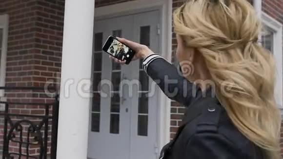 一个美丽的金发女孩的侧面肖像她用智能手机上网在一个郊区的家中拍摄自拍照片视频的预览图