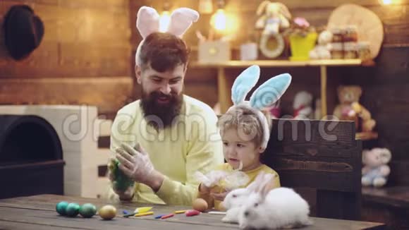父亲和儿子给复活节彩蛋上色可爱的小男孩戴着兔子耳朵木制背景的复活节彩蛋快乐视频的预览图