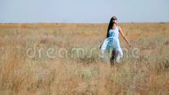 穿着蓝色连衣裙的年轻漂亮女孩喜欢花草原草地上的年轻女子草原阳光明媚的草地视频的预览图
