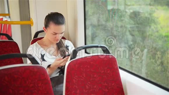 年轻女孩在乘坐公共汽车时使用智能手机美丽的女人在城市交通中使用她的手机视频的预览图