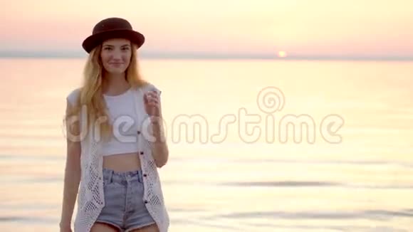 戴着帽子的漂亮年轻女子在海洋和金色夕阳的背景上摆姿势视频的预览图