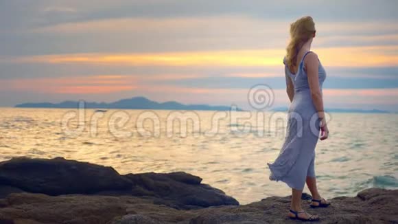 年轻女子走在岩石上欣赏日落美景溅起浪花视频的预览图