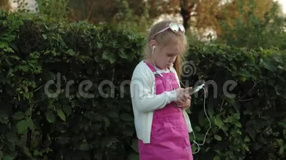 一个穿着粉红色裙子的小女孩坐在台阶上的公园里戴着耳机享受着智能手机视频的预览图