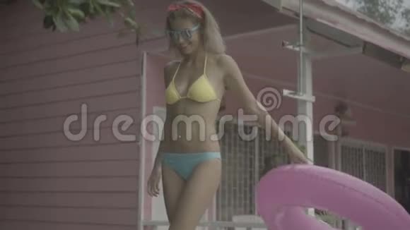 穿着比基尼戴着太阳镜带着粉色充气戒指走着的漂亮年轻快乐的女人在粉色房子的背景下被隔离开来视频的预览图