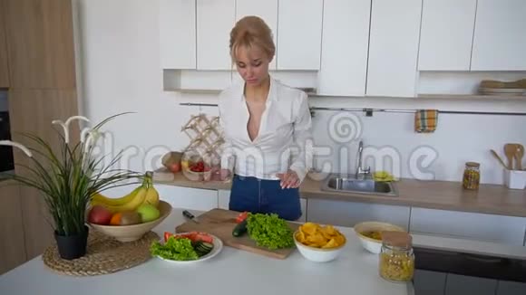 苗条的金发女人站在现代餐桌前准备好健康的早餐传播蔬菜视频的预览图