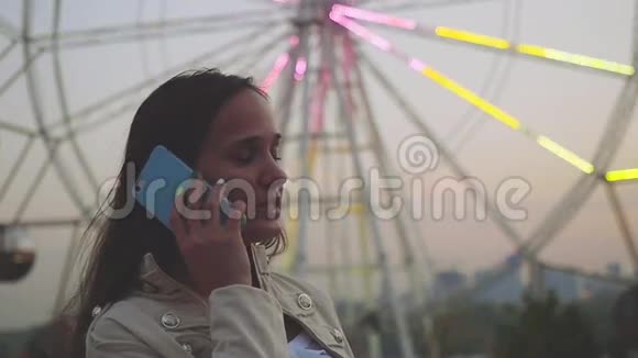 一位年轻的女士在一个景点公园的街机旁在慢动作的摩天轮发光的背景下用智能手机交谈视频的预览图