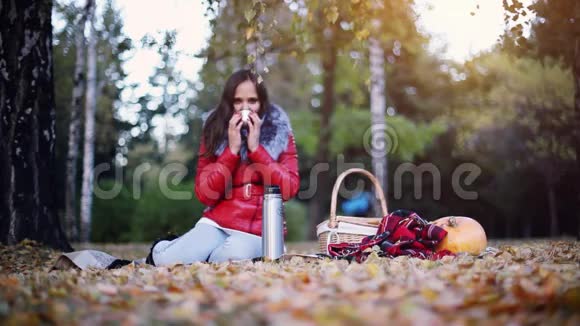 年轻漂亮的女人坐着野餐在秋天公园里喝热茶坐在地毯上的女孩视频的预览图
