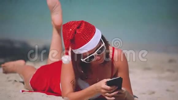 圣诞老人戴着红色圣诞帽的年轻微笑女子在海边的沙滩上拍照自画像视频的预览图