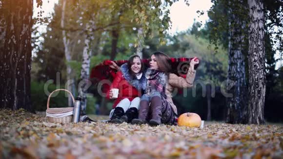 秋天公园里年轻的黑发女人坐在野餐的毯子里喝着热茶落叶上的女孩视频的预览图
