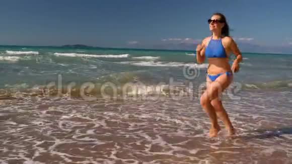 海滩比基尼女郎无忧无虑地沿着海滩上的水奔跑希腊科孚海岸风景如画视频的预览图