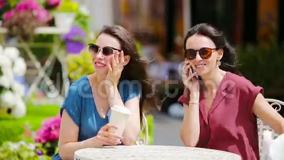 两个女孩在户外咖啡馆用智能手机两个女人在买完包后坐在露天咖啡馆视频的预览图