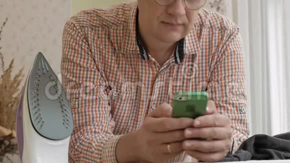一个男人在他家的熨衣板上抚摸他的衬衫用智能手机视频的预览图
